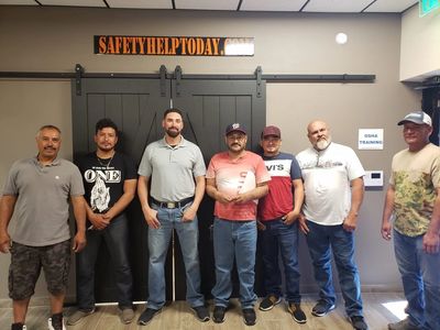 Seven men standing together inside a room with a safety sign above.