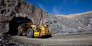 A large yellow mining truck near a rocky tunnel entrance under a clear blue sky.