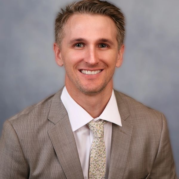 Smiling man in a beige suit and patterned tie against a gray background.