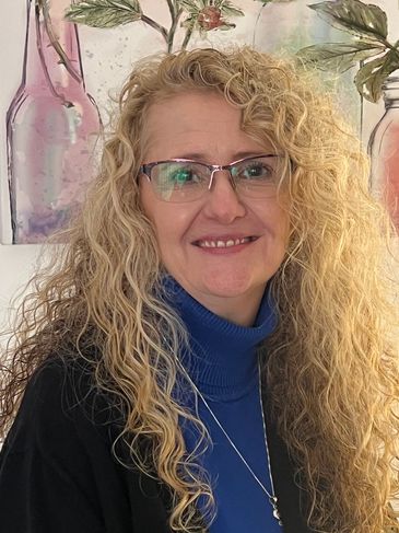 Smiling woman with curly blonde hair and glasses wearing a blue turtleneck.