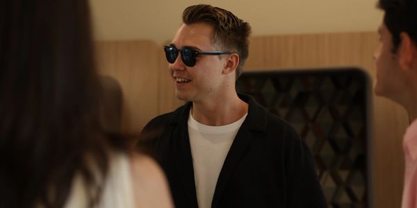 A man wearing sunglasses smiles while talking to others indoors.
