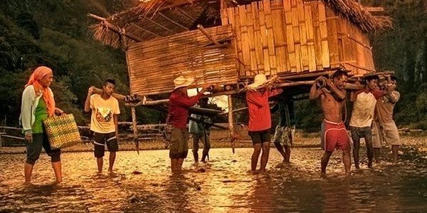 People carrying a bamboo hut through shallow water during sunset.