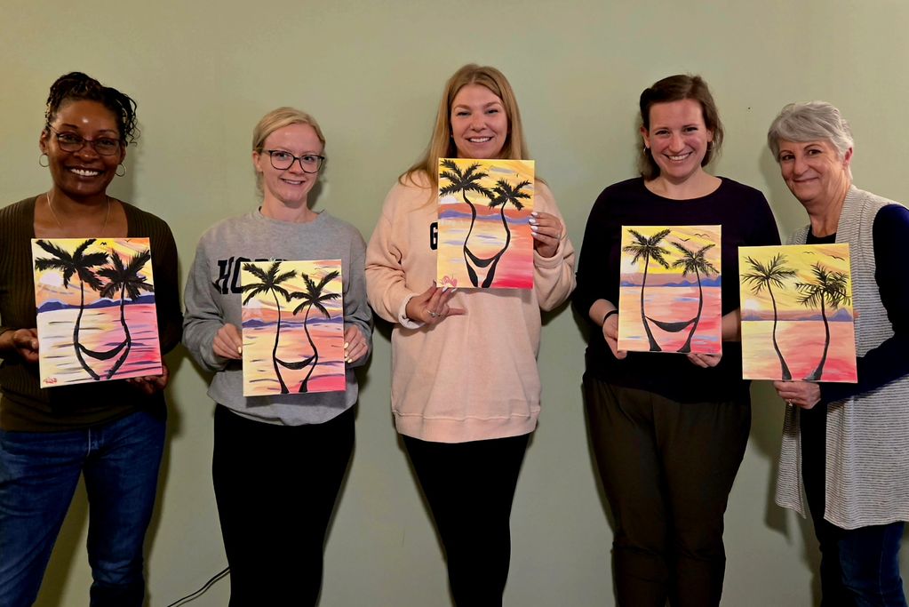 Five women proudly display their sunset palm tree paintings in a group photo.