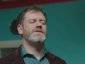 Man with closed eyes and beard in a teal room.