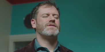 Man with closed eyes and beard in a teal room.