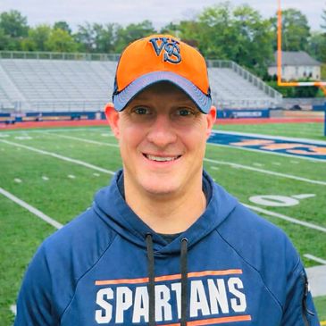 Man wearing a Spartans hoodie and a baseball cap on a football field.
