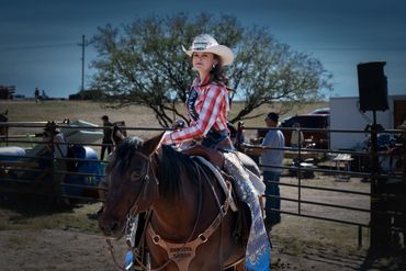 Young rodeo princess in western attire rides a horse confidently at an outdoor event.