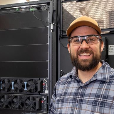 Smiling man in safety glasses standing next to technical equipment rack.