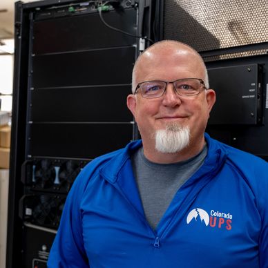 Man wearing glasses and a blue Colorado UPS jacket smiling indoors.