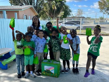 A group of children and two adults smiling outdoors with green gift bags.