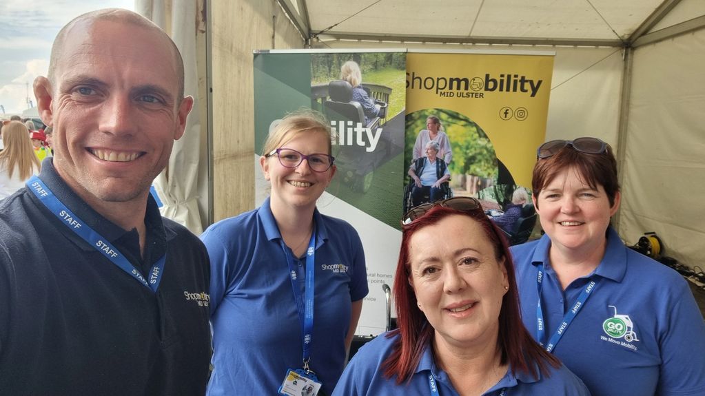 Four smiling staff members in blue shirts at a Shopmobility Mid Ulster booth.