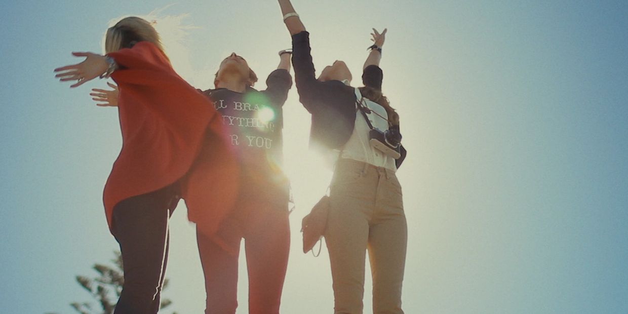 Three women joyfully reaching up towards the sky with sunlight behind them.