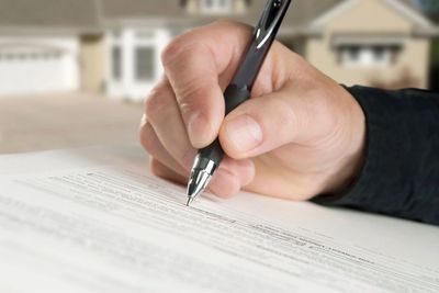 Close-up of hand signing a document with pen, blurred house in the background