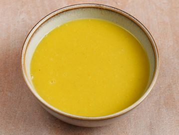 A bowl of smooth yellow soup on a beige surface.