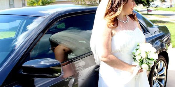 Bride in white dress holding bouquet next to black car.