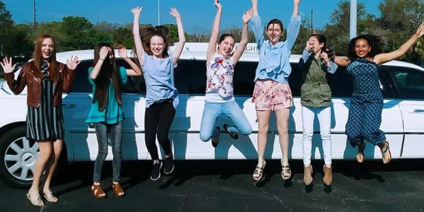 Seven girls jumping joyfully in front of a white limousine.