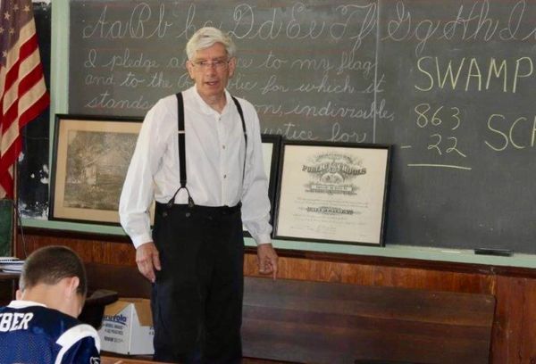 Elderly man teaching in a vintage classroom with blackboard and American flag.