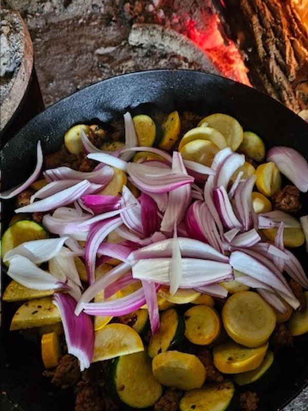 Sliced onions and zucchini cooking in a pan over an open flame.