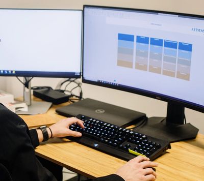 Two computer monitors side-by-side on a desk with a keyboard, someone's hands are typing on it. 