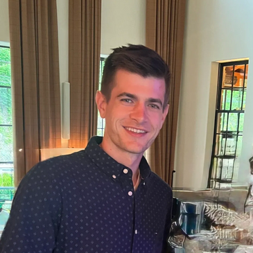 A young man smiling, standing indoors in a stylish room with stone walls and large windows.