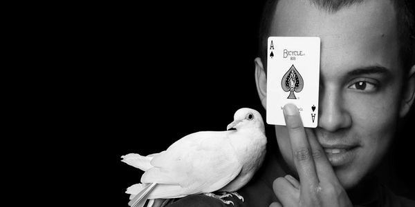Man holding ace of spades card over one eye with a white pigeon on his shoulder.