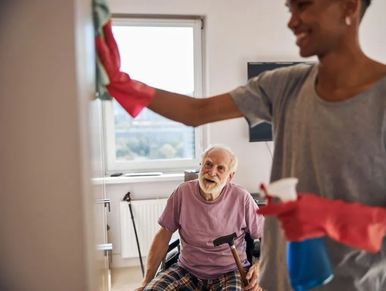 A caregiver cleans while an elderly man watches from his wheelchair.