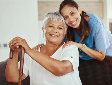 Smiling elderly woman with walking cane and caregiver in blue scrubs sharing a warm moment.