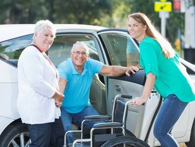A man with a wheelchair and two women by a car outdoors.