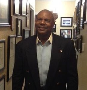 Man in a dark blazer smiling in a hallway filled with framed pictures.