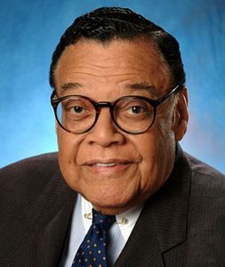 Portrait of an elderly man in glasses and a suit against a blue background.