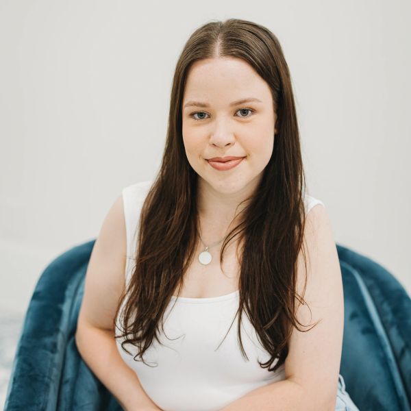 Young woman with long brown hair sitting on a blue chair, smiling gently.