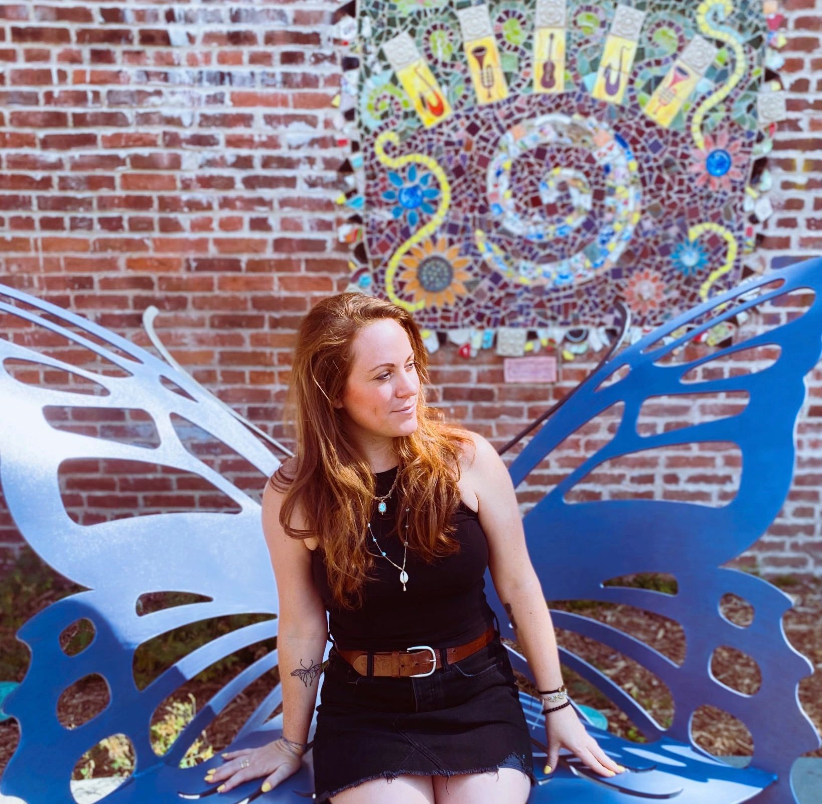Woman sitting on a blue butterfly-shaped bench against a brick wall with a colorful mosaic.