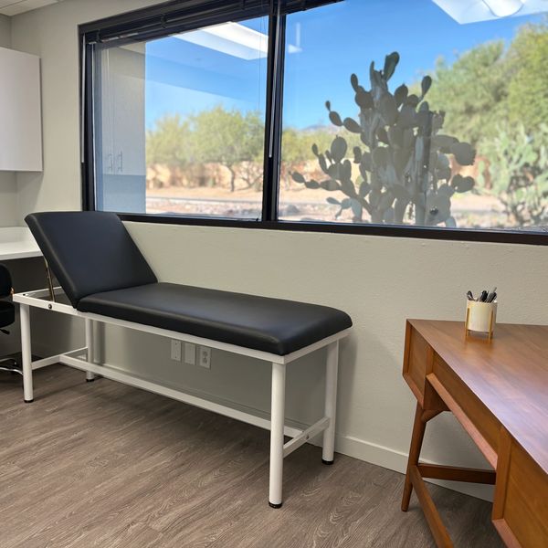 A clean medical exam room with a black exam table and a wooden desk.
