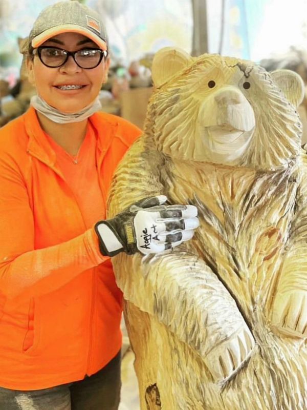 Woman in orange jacket posing with a carved wooden bear statue.