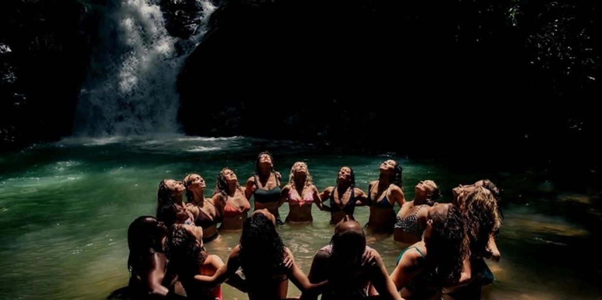 Women in bikinis standing in a circle in a waterfall pool, heads tilted back.