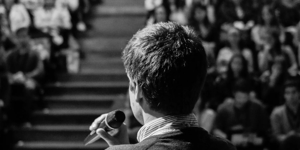 Man speaking into a microphone to an audience in black and white.