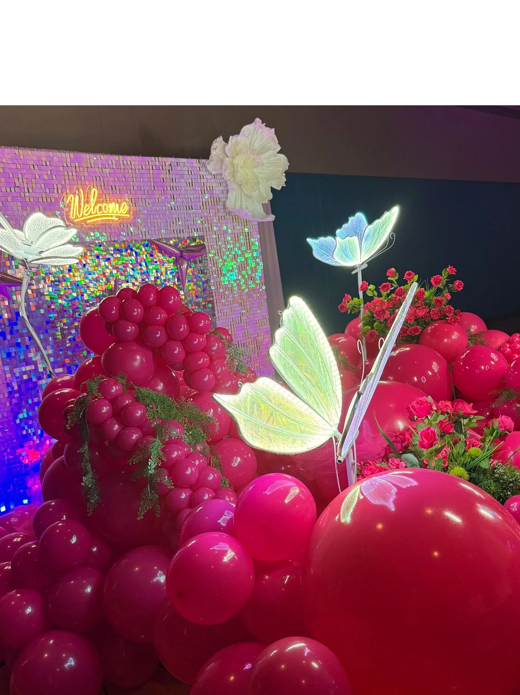 A vibrant balloon and flower decoration with illuminated butterflies and a glowing 'Welcome' sign.
