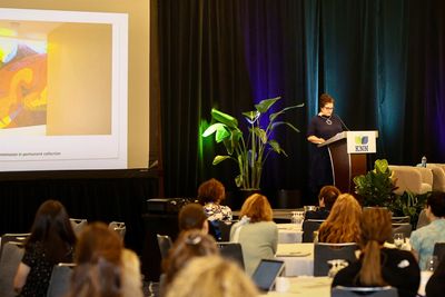 A woman speaks at a conference podium with an audience and presentation screen.