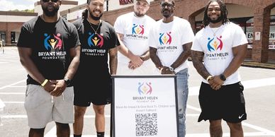 Five men wearing Bryant Helen Foundation shirts stand outdoors with a sign.