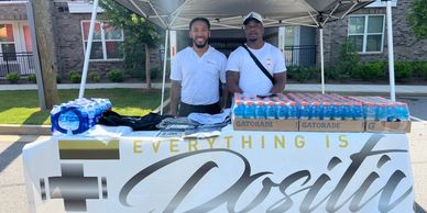 Two men stand behind a table with water bottles and snacks under a canopy.