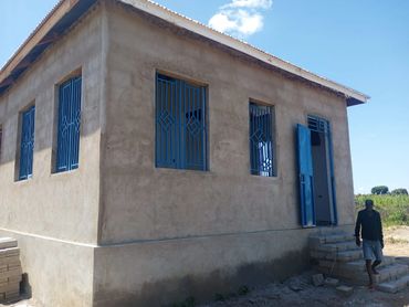 A small unfinished concrete house with blue windows and a man walking nearby.