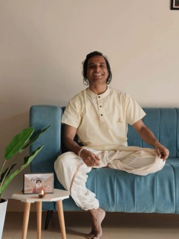 Man in traditional attire sitting cross-legged on a blue sofa, smiling.