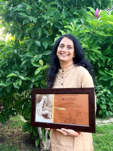 Woman smiling outdoors holding a framed certificate with greenery in the background.