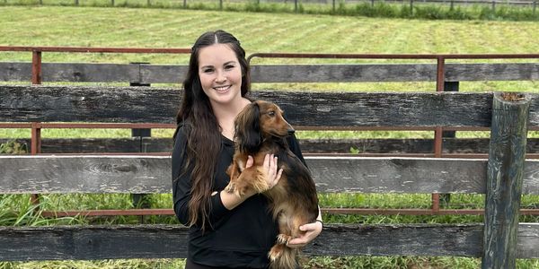 Smiling woman holding a dachshund in front of a wooden fence.