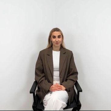 Woman in brown blazer sitting on black chair against white wall.
