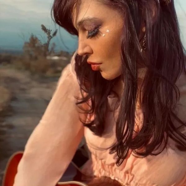 Woman with long hair and star stickers on her cheek, wearing a peach dress outdoors.