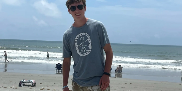 Young man wearing sunglasses standing on a sandy beach with ocean waves in the background.