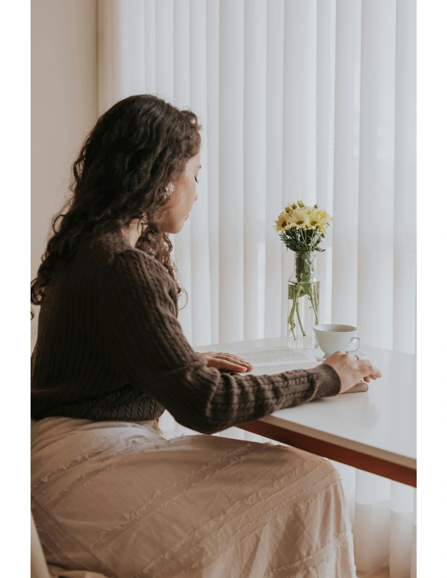 Woman reading by a window with flowers and a cup of tea.