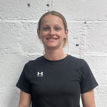 Woman smiling in a black Under Armour t-shirt against a white textured wall.