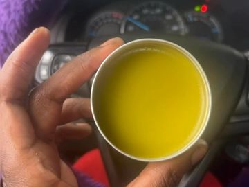 Hand holding a cup of yellow liquid inside a car with a purple steering wheel cover.